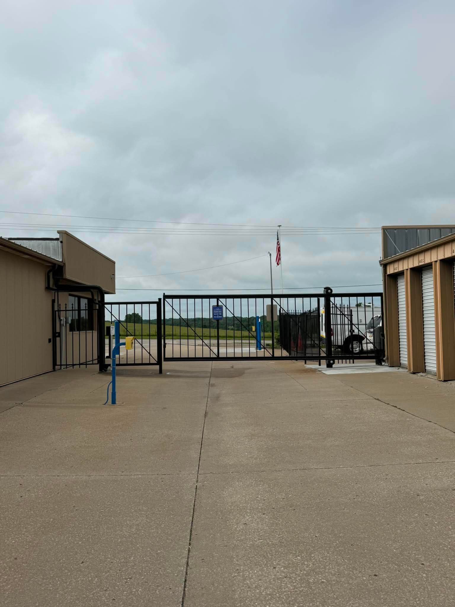 Black metal gate at a storage facility entrance between tan buildings under a cloudy sky.