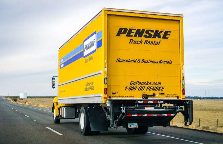 Yellow Penske rental truck driving on a highway through a rural landscape under cloudy skies.