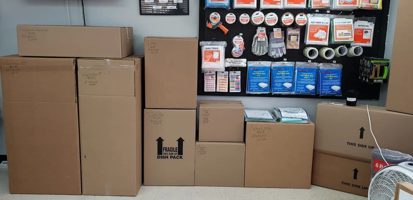 Stacked cardboard moving boxes of various sizes next to a wall of packing supplies.
