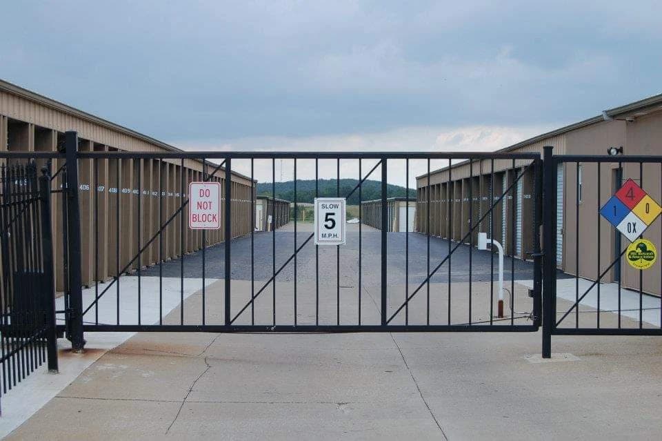 Closed black metal gate at a storage facility entrance with speed limit and warning signs.