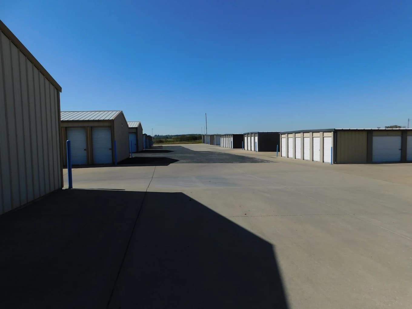 Tan self-storage units with white roll-up doors line a paved lot under a clear blue sky.